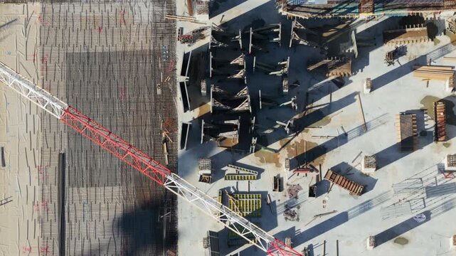 Aerial top-down view showing progress at a skyscraper construction site, with reinforced concrete foundations, steel rebar grids, formwork, and construction materials casting long shadows