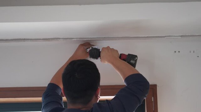 Man using cordless drill to install fixture on white ceiling near window frame in home renovation