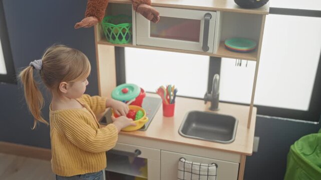 Girl playing with toy dishes at a play kitchen reaches for cup by sink in a building playroom; curiosity discovery.