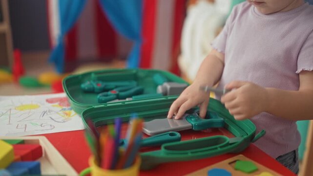 Girl playing with green toy medical kit, holding plastic pliers with bare hands in preschool classroom; playful curiosity.