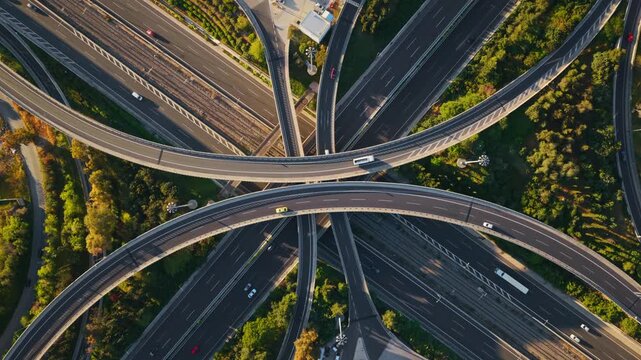 Gradual aerial zoom over a complex multilevel highway interchange with traffic moving along the various lanes, viaducts, and overpasses, creating a symmetrical pattern surrounded by green areas