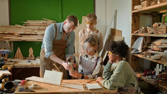 Gen Z craftsman teaching teenage girl to burn wood using gas torch during carpentry class, with two diverse boys watching