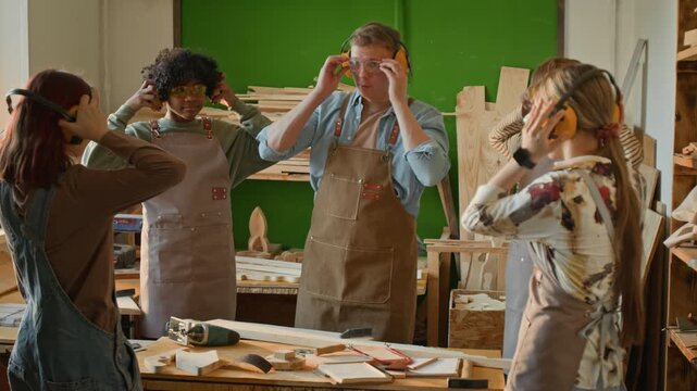 Gen Z male teacher asking teenage group to put on safety earmuffs and showing them how to work with jigsaw during woodworking class