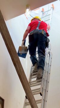 Male construction worker in a hard hat and red t-shirt climbing an extension ladder on a building site.