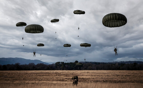 Military paratroopers in green parachutes descend toward a vast dry field under a moody, overcast sky, representing strategic deployment, tactical training, and airborne operations.