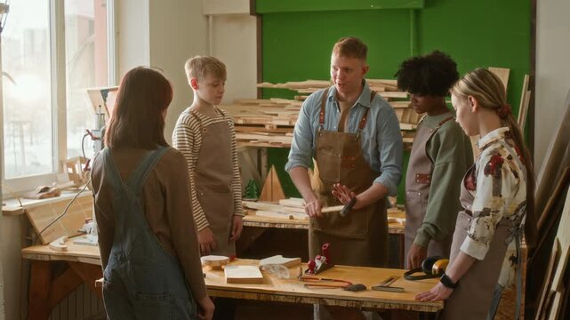 Young craftsman instructing teenage group on how to use hammer and handing it to boy during woodworking masterclass
