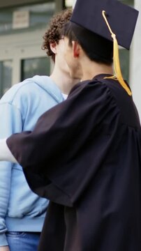Vertical video: Coming grad gown hugging friend by door saying cheers and reaching cell, copy space
