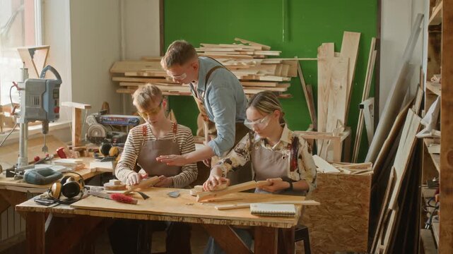 Two teenagers polishing wooden details under supervision of gen Z male carpenter conducting masterclass in his workshop