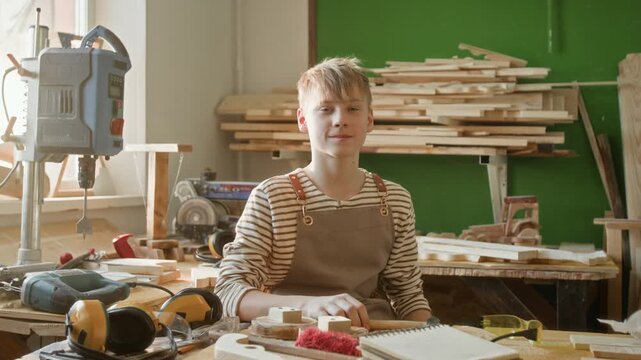 Portrait of blond Caucasian teenage boy taking off safety goggles and smiling at camera during craft lesson in carpentry workshop