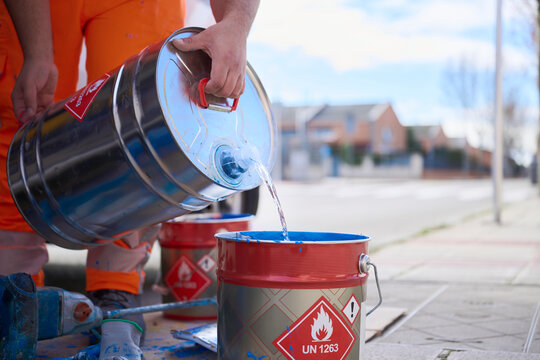 Worker pours liquid solvent into blue road marking paint in a bucket preparing materials for application on asphalt while managing flammable substances safely.