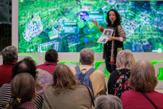 Group of seniors listening to urban planning presentation, speaker gestures toward large digital map, community engagement, public meeting.