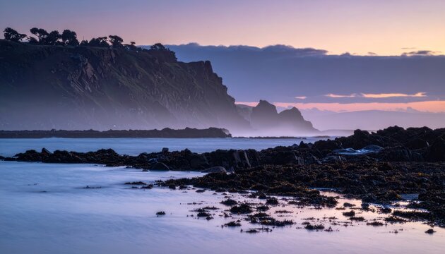 A serene coastal landscape showcases a dramatic cliff face bathed in the soft hues of twilight, with gentle waves lapping against a rocky shoreline.