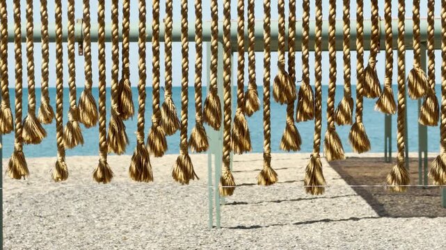 Rustic ropes with frayed tassels sway gently in the breeze. Their coarse textures and the calming view of the blue sea create a deeply relaxing, tropical feeling of absolute serenity.