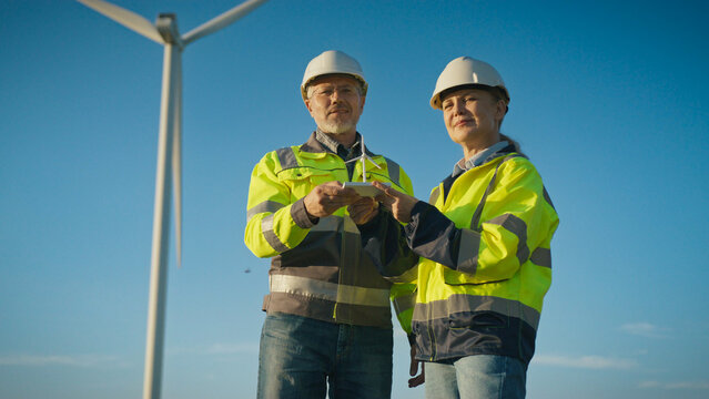 Two workers in helmets and reflective jackets holding miniature wind turbine together. Man and woman standing in field with large turbine behind. Engineers presenting renewable energy model.
