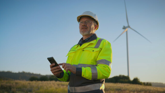 Worker standing in field near wind turbine and smiling while checking information on device. Renewable energy inspection