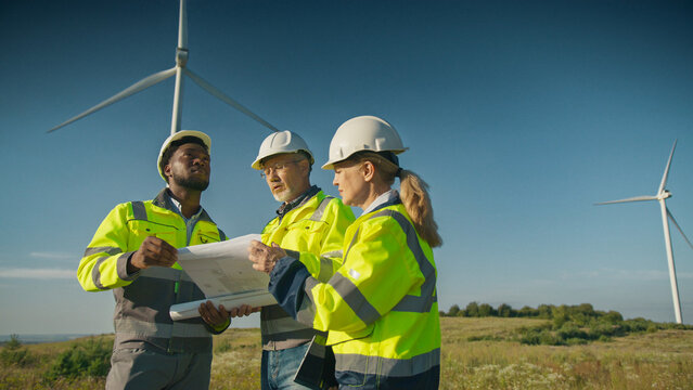 Three engineers in helmets and reflective jackets standing in field near turbines. Workers holding blueprint and tablet. Team deciding about building new wind turbine location under clear sky.
