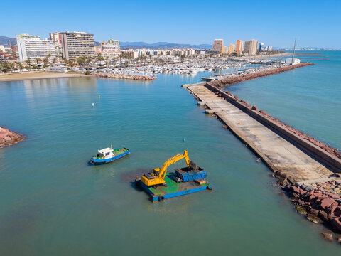 Yellow excavator on floating barge performs harbor dredging near a breakwater jetty, with a marina, palm trees, and coastal city skyline visible in the background