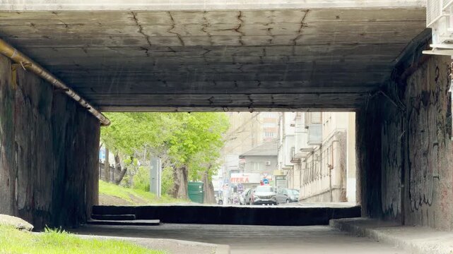 Dark rain pours through a gloomy, weathered concrete underpass. The wet, dreary mood contrasts with the bright green trees and quiet city street seen just ahead.