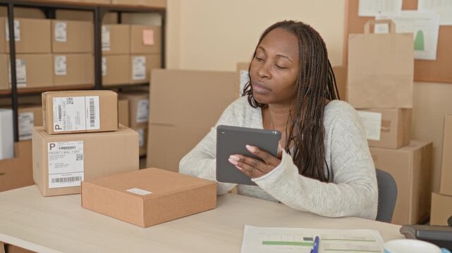 Woman holding tablet among stacked shipping boxes in a building workspace checking parcels and labels with a thoughtful expression; small business optimism.