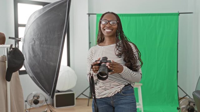Woman photographer holding camera and lens, smiling and giving thumbs up in studio with green backdrop and softbox; confidence creativity.