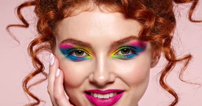 Stylish woman with rainbow eyeshadow makeup and curly hair posing elegantly on pink background
