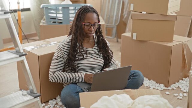 Woman typing on laptop while sitting crosslegged among packed moving boxes and scattered packing peanuts in building interior; focus transition.