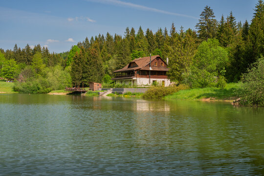View of Krpacovo reservoir and a traditional wooden lodge surrounded by lush green pine forests and calm water reflections under a clear sky Dolna Lehota, Banskobystricky kraj, Slovakia.