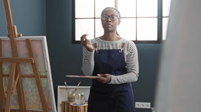 Woman painter holds palette and brush and touches face while evaluating canvas in studio; contemplation creative process.