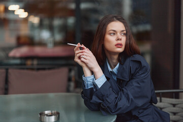 Fototapeta premium Young woman with long brown hair wearing a dark coat smokes a cigarette while sitting at a glass table in a dimly lit indoor cafe.