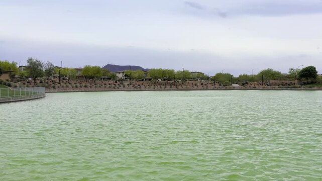View of a choppy lake with green waves on a windy, overcast day. In the distance, suburban house roofs and cars moving along the road are visible. Cool atmosphere of a cloudy day in a Nevada park.