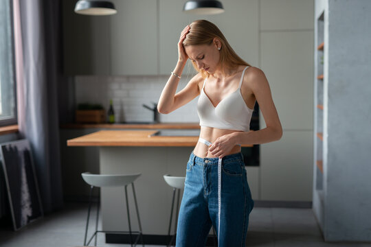 Young woman in a modern kitchen looks distressed, holding her head while measuring her waist with a tape, reflecting anxiety, body image struggle and dieting pressure