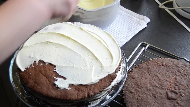 Close-up of female hands spreading smooth creamy frosting on chocolate sponge cake layers while preparing a homemade layered dessert.