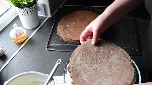 Close-up of female hands spreading smooth creamy frosting on chocolate sponge cake layers while preparing a homemade layered dessert.