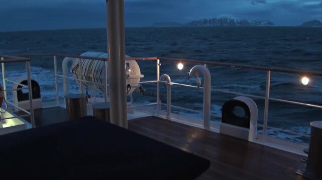 Cruising the vast Arctic Ocean off Norway at twilight. Warm deck lights reflect on dark, undulating waves, while majestic mountains grace the distant horizon under a deep blue sky.