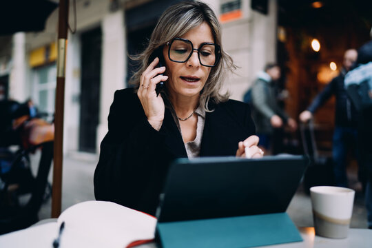 Caucasian middle aged woman in glasses phoning and using tablet with stylus on terrace, busy female entrepreneur multitasking with mobile phone and digital device, remote work and data analysis.