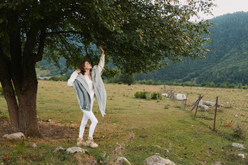 Naklejka premium A woman in a light cardigan dances beside a tree in a rural field, capturing a carefree outdoor moment and a sense of freedom, a lifestyle scene of wellness and nature.