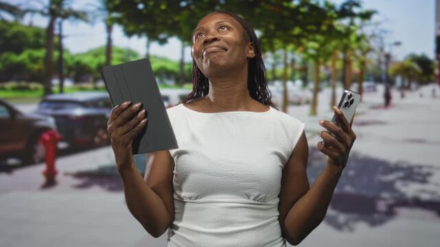 Young african american woman holding tablet and smartphone, eyes closed and head tilted back in a pleased gesture on a sunny street downtown; contentment.
