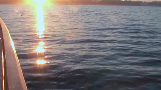 Witness the breathtaking sunset over the Arctic Ocean from a boat near Norway. Golden sunbeams create a sparkling path on the calm, undulating waters. A tranquil and beautiful evening unfolds.
