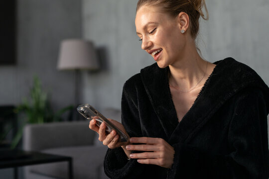 Young woman in a cozy black robe laughing at her smartphone while relaxing at home, enjoying morning scrolling and connecting with friends through social apps