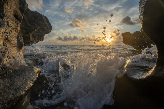 View of ocean waves crashing against dark coastal rocks under a golden sunrise in Florida, United States.