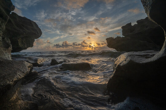 View of the sun rising over the ocean waves framed by dark coastal rock formations in Florida, United States.