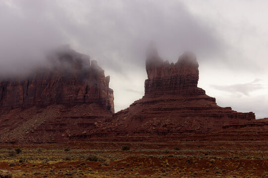 View of majestic red sandstone buttes partially shrouded in thick fog and low clouds under an overcast sky in Oljato-Monument Valley, Arizona, United States.