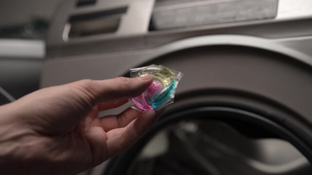 Person hand tossing colorful laundry detergent pod capsule into washing machine drum filled with dirty clothes, getting ready for fresh wash cycle and household chores, close-up, slow motion.