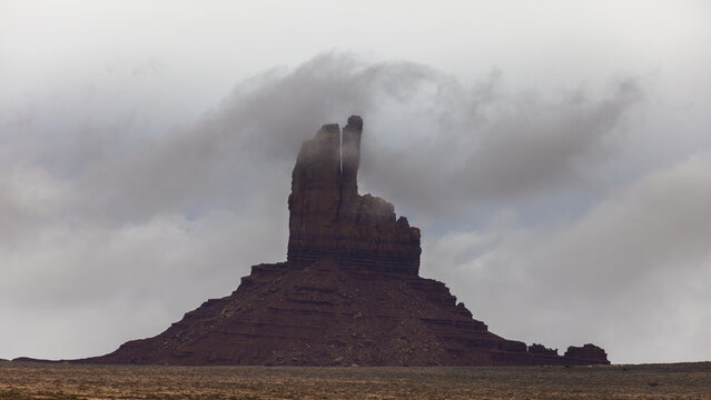 View of the iconic West Mitten Butte sandstone formation partially shrouded in low-hanging clouds and mist under a dramatic overcast sky in Oljato-Monument Valley, Arizona, United States.
