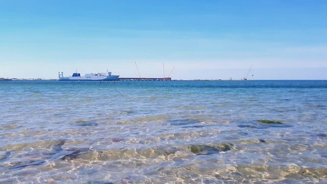 Seascape on Baltic sea coast with calm water surface of clean turquoise coastal surf waves over sand bottom and large ferry boat moored near berth at seaport quay under clear blue sky at sunny day