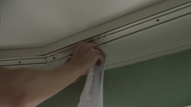 Close-up of unrecognizable person installing white sheer curtain by attaching hooks to ceiling mounted track system, performing common home improvement, interior decoration task in room, slow motion.