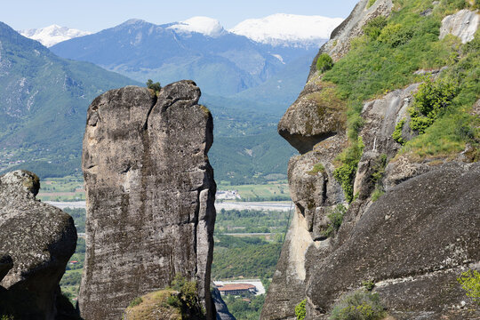 monolithic rock pillars of meteora stand tall against a backdrop of snow-capped pindus mountains and a lush green valley in greece under a clear blue sky