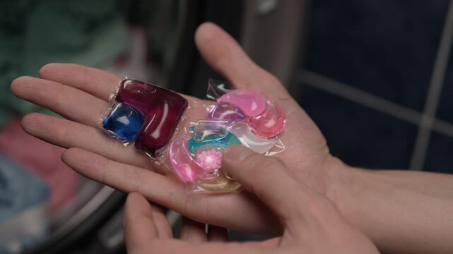 Close-up of laundry day routine showing person holding colorful liquid laundry detergent pods in their palm and then tossing into modern washing machine filled with dirty clothes, slow motion.