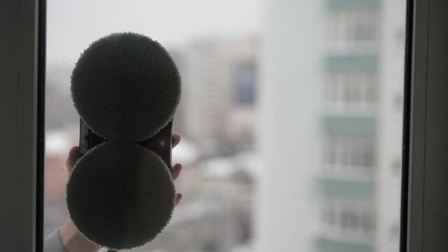 Person attaching modern robotic window cleaner to glass. Robotic washer with rotating microfiber pads cleaning dirty window in apartment. Modern smart home technology assisting with household chores.