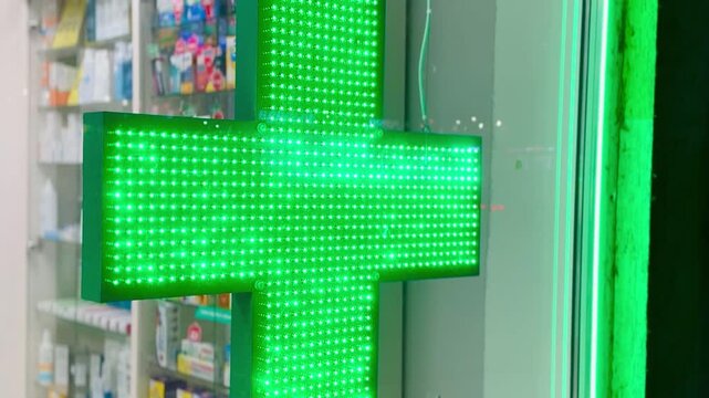 Green led pharmacy cross sign hanging, vibrant diode matrix pulses across suspended fixture, glass storefront reflection, clinical